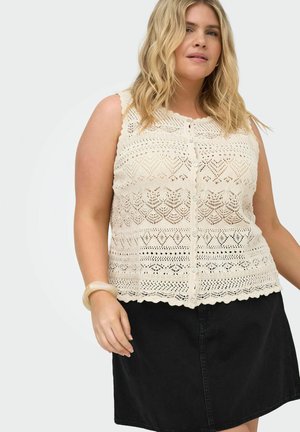 Blonde Frau trägt ein ärmelloses cremefarbenes Häkel-Button-Top und einen schwarzen Jeansrock, posiert mit einem beigen Armband am Handgelenk.