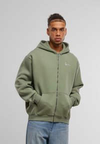 Green zip-up hoodie made of soft fabric, featuring a kangaroo pocket, ribbed cuffs and hem, with a small logo on the chest.