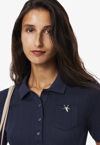 Mørkeblå polo skjorte med struktureret stof, der har en lomme og en dekorativ nål med en figur med grønne accenter.