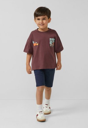 Młody chłopiec idzie, ubrany w bordowy t-shirt z motywem ananasów, granatowe krótkie spodenki, białe skarpetki i białe buty sportowe z czerwonymi akcentami.