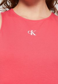 Žena nosí bezrukávový korálově růžový top s malým bílým logem "ck" Calvin Klein na přední části u výstřihu.