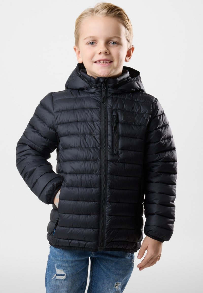 Giacca nera imbottita con chiusura a zip, cappuccio regolabile e polsini elasticizzati. Presenta una tasca con zip sul davanti e orlo a coste. Indossata su jeans di denim.