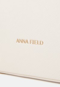 Cream-colored textured surface with a subtle diagonal pattern. Features embossed gold brand name "ANNA FIELD" at the center.