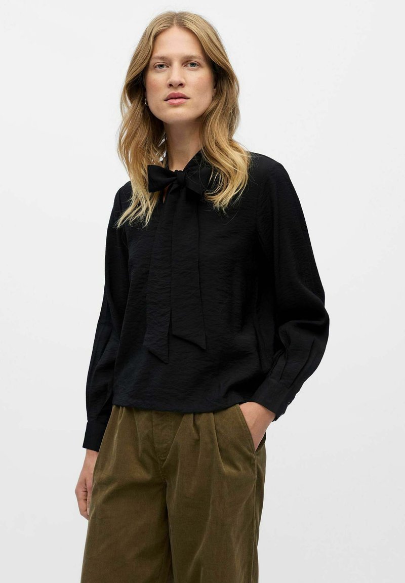 Blouse noire avec une finition texturée, nœud papillon au niveau du col et manches longues, associée à un pantalon en velours côtelé vert olive.