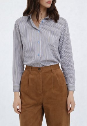 Donna che indossa una camicia a righe verticali blu e bianche infilata in pantaloni in camoscio marrone a vita alta, in piedi con le braccia rilassate.