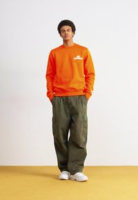 Spilgti oranžs krekls ar priekšējo logotipu, apvienots ar brīvi pieguļošām olīvzaļām kravas biksēm. Baltas kedas noslēdz apģērbu.