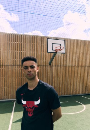 T-shirt Nike noir avec un graphique de taureau rouge. En arrière-plan, des murs en bois et un panier de basket ; un terrain vert avec des marquages blancs. Ciel bleu au-dessus.