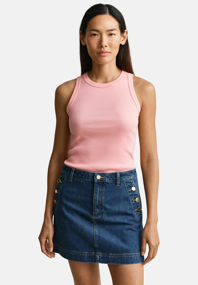 Femme aux longs cheveux foncés portant un haut rose côtelé sans manches et une mini-jupe en jean bleue avec des boutons dorés sur les poches.