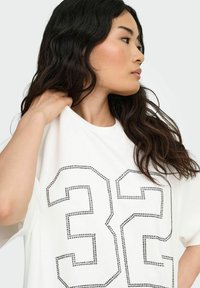 Maglietta bianca a maniche corte con il numero "32" contornato da borchie nere. Tessuto morbido con una vestibilità comoda, presenta un collo a giro.