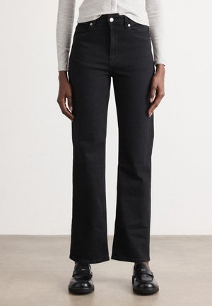 Personne portant un jean noir droit taille haute, un haut gris clair à manches longues et des mocassins noirs, debout sur un sol neutre contre un mur blanc.