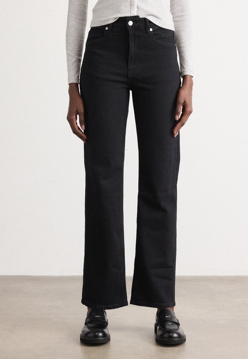 Personne portant un jean noir droit taille haute, un haut gris clair à manches longues et des mocassins noirs, debout sur un sol neutre contre un mur blanc.