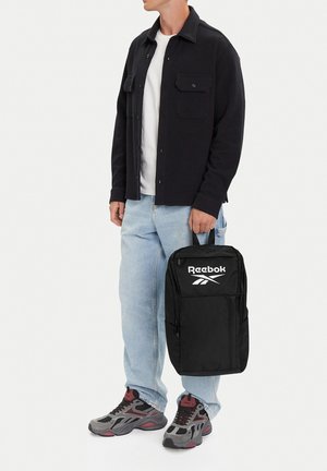 Mann trägt schwarze Jacke mit Knöpfen, weißes Hemd, hellblaue Jeans, graue und rote Sneakers, hält schwarzen Reebok-Rucksack am Griff.