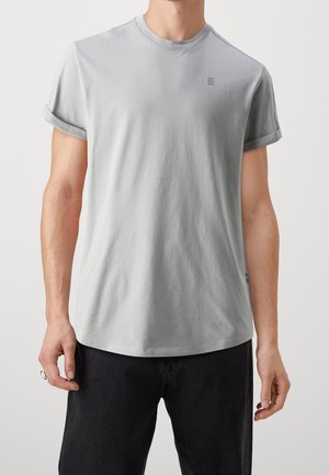 Mężczyzna w jasnoszarej koszulce z krótkim rękawem i dekoltem w serek, z podwiniętymi mankietami, oraz czarnych spodniach, stojący na jednolitym tle.