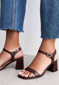 Brązowe skórzane sandały na obcasie o fakturze krokodyla, z odkrytymi palcami i paskiem na kostkę z srebrną klamrą, w zestawie z przyciętymi dżinsami.