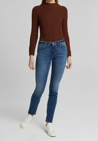 Jersey de punto marrón con cuello alto, combinado con vaqueros azul ajustados y zapatillas blancas. El jersey presenta un patrón texturizado.