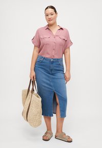 Chemise boutonnière rose clair à manches courtes, jupe en denim bleu taille haute avec fente avant, sac fourre-tout en paille et sandales à glissière marron.