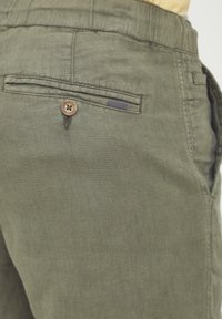 Pantaloni in lino verde oliva con una tasca posteriore con bottone. Presentano una texture liscia e una piccola linguetta decorativa sopra la tasca.