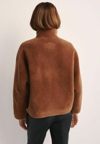 Brązowa polarowa kurtka z wysokim kołnierzem, luźnym krojem i teksturowaną powierzchnią. Brak widocznych elementów metalowych lub akcentów na widoku z tyłu.