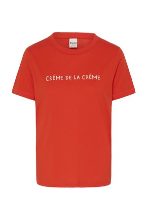 Sarkans kokvilnas T-krekls ar īsām piedurknēm un apaļu kaklu. Priekšpusē centrā redzams baltais teksts "CRÉME DE LA CRÉME".