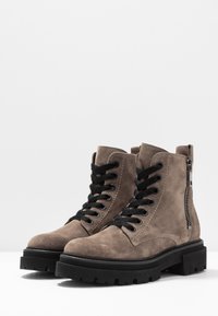 Bottines en daim marron clair avec lacets noirs, fermeture éclair sur le côté, semelle en caoutchouc noir épaisse et détails de couture contrastants.