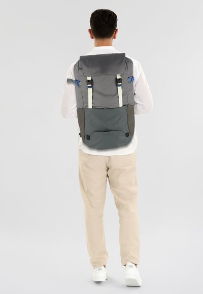 Mochila de tela gris y gris oscuro, con correas ajustables, cierre de solapa y detalles en hardware azul. Diseño simple con un compartimento espacioso.