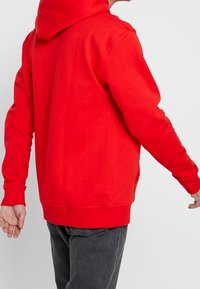 Persona con felpa con cappuccio rossa accesa, vista di spalle, abbinata a jeans grigi scuri, in piedi contro uno sfondo chiaro e semplice.