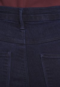 Jeans de mezclilla oscura con una textura suave, diseño ajustado y dos bolsillos traseros con costuras detalladas. La cinturilla se pliega para ajustar el ajuste.