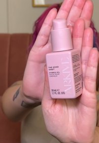 Bouteille rectangulaire rose étiquetée "L'Huile Hybride de Bonnes Habitudes", avec un capuchon à pompe, contenant 50 mL de produit. Design minimaliste, finition brillante.