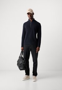 Mørkeblå ribbet sweater med kvart lynlås, sorte jeans, beige kasket med logo, beige sneakers og en sort lædertaske, stående på en hvid baggrund.