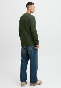Groene gebreide trui met een geribde zoom, gedragen met lichtblauwe loszittende jeans en beige laarzen, gezien van achteren.
