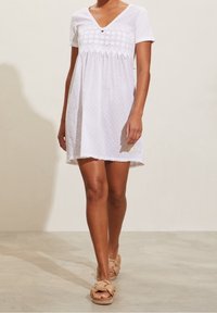 Vit kort klänning med V-ringning, spetsinlägg och prickigt mönster. Flödande design med scalloped nederkant, i sällskap av beige flätade sandaler.