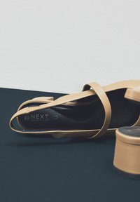 Bež cipele s visokom petom minimalističkog dizajna, s tankim trakama i zaobljenim vrhom. Crna unutrašnjost s markom "NEXT MOTIONFLEX".