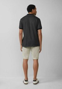 Schwarzes Polohemd mit kurzen Ärmeln, kombiniert mit hellbeigen Shorts und weißen Schuhen. Das Polohemd hat einen Kragen und kontrastierende Bündchen.