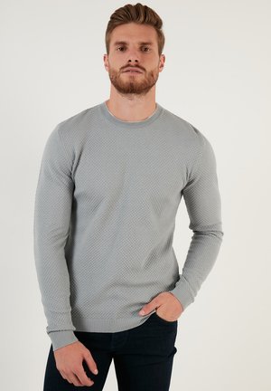Pull en tricot gris clair avec un motif texturé, encolure ronde et poignets côtelés. Associé à un jean bleu foncé.