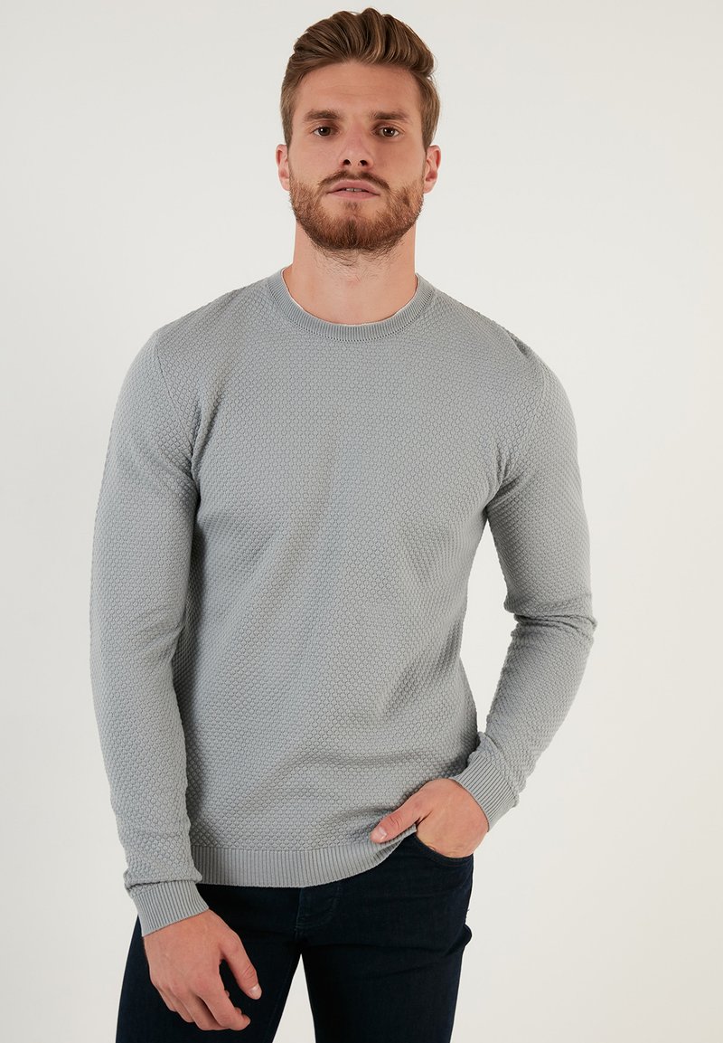 Maglione lavorato a maglia grigio chiaro con un motivo testurizzato, scollo rotondo e polsini a costine. Abbinato a jeans blu scuro.