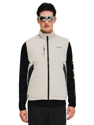 Mand iført lys beige ærmeløs lynlåsvest over sort langærmet skjorte, med reflekterende solbriller, stående mod en hvid baggrund.