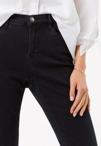 Jeans denim biru tua dengan pinggang tinggi, saku depan yang terlihat, dan kancing perak. Dipadukan dengan kemeja putih lengan panjang.