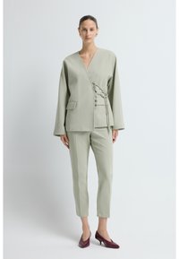 Vrouw staat in een lichtgrijze kostuum met een wikkeljack en broek tot de enkels, gedragen met donkerrode schoenen met lage hakken en gouden oorbellen.