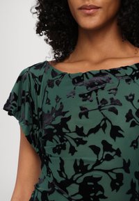 Blouse à motif floral vert en tissu doux, avec des accents veloutés noirs et un large col, comportant des détails froncés sur les côtés.