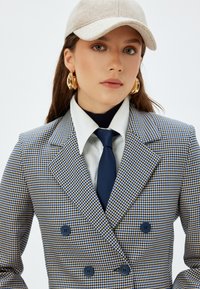 Dubbelzijdige jas in blauw, geel en witte houndstooth patroon, met grote blauwe knopen, een wit shirt en een donkerblauwe das.