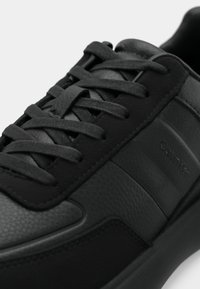 Μαύρα δερμάτινα sneakers χαμηλής κορυφής με επίπεδα κορδόνια, εξαιρετική υφή, διακριτική επωνυμία και λείο αντίθετο πάνελ στο πλάι.