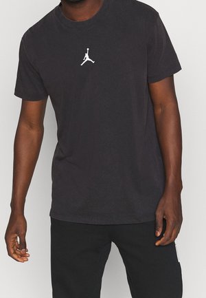 Homme portant un t-shirt noir avec une petite silhouette blanche d’un joueur de basket en plein saut, associé à un pantalon noir.