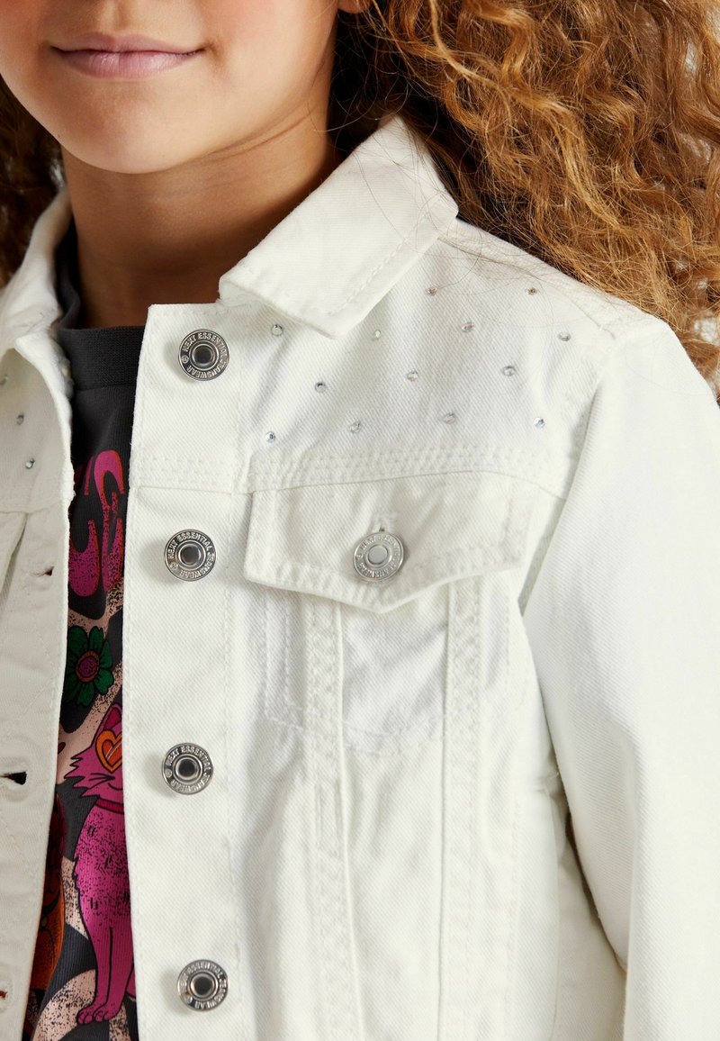 Enfant aux cheveux bouclés portant une veste en jean blanche avec de petits strass et des boutons argentés sur un t-shirt graphique coloré.