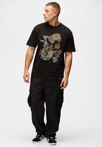 Sort bomuld t-shirt med et farverigt "Space Jam" tegnefilm grafik på forsiden. Båret med løstsiddende sorte cargo-bukser og sorte sko.
