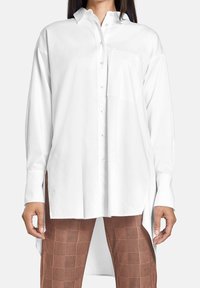 Chemise blanche à manches longues avec boutons, col et poche poitrine, présentant un design fluide avec des fentes latérales et un tissu léger.