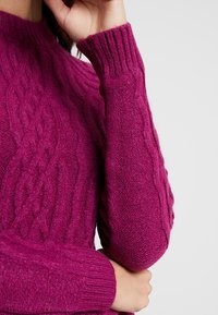 Fuchsia gestrickter Pullover mit geripptem Halsausschnitt und Bündchen, der ein strukturiertes Zopfmuster entlang der Ärmel und des Körpers aufweist.
