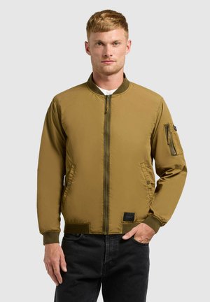 Szőke férfi khaki bomber dzsekiben, elöl cipzárral, két oldalzsebbel és egy cipzáras zsebbel a bal ujján, fekete farmernadrággal párosítva.