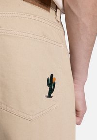 Pantalones de mezclilla beige con un bordado de cactus verde en el bolsillo trasero, con un tejido texturizado y detalles de costura en contraste.