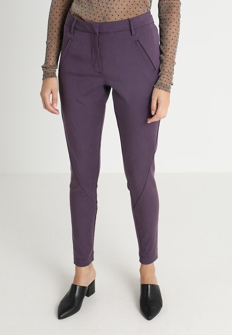 Femme portant un pantalon violet ajusté avec des poches avant inclinées, un haut manches longues en tulle à pois, et des mules noires pointues à talons blocs.