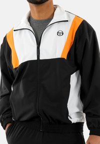 Chaqueta con cremallera en negro, blanco y naranja, fabricada con un tejido ligero; cuenta con un cuello alzado y puños y dobladillo elásticos. Logotipo en el pecho.